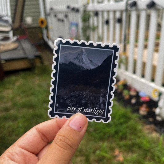 Sticker in shape of postage stamp with a mountain city and the words "city of starlight" inspired by ACOTAR's Velaris