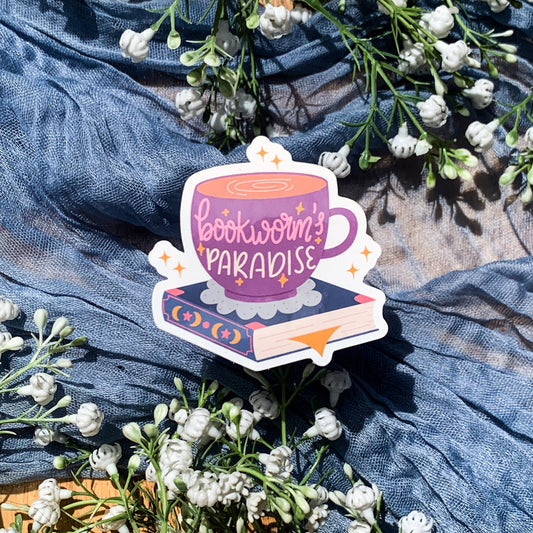 A bookish aesthetic booktok sticker on a purple small cup of coffee on top of a book with the quote "bookworm's paradise" on the front of the cup surrounded by yellow sparkles. 