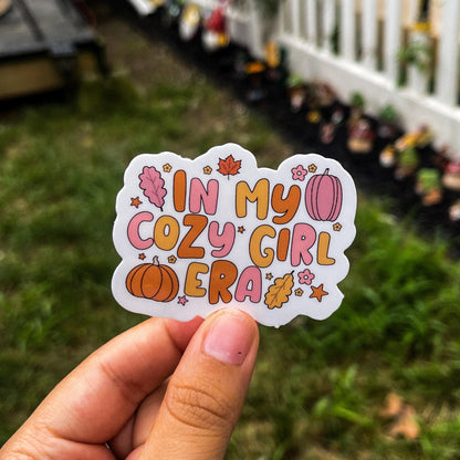 Aesthetic fall sticker with the quote "in my cozy girl era" surrounded by leaves, pumpkins, flowers, and stars. the overall color scheme is orange, pink, and yellow alternating between each object.