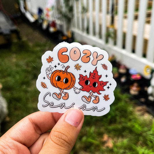 A cozy fall inspired sticker featuring a cartoon pumpkin and leaf holding hands and the words "cozy season".