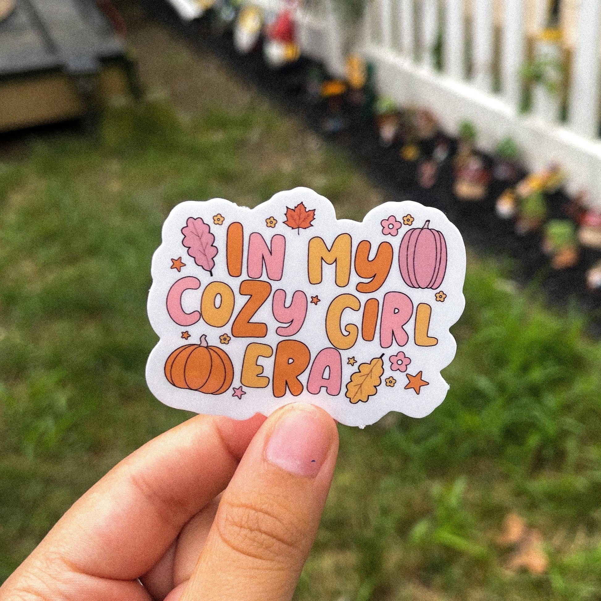 Aesthetic fall sticker with the quote "in my cozy girl era" surrounded by leaves, pumpkins, flowers, and stars. the overall color scheme is orange, pink, and yellow alternating between each object.