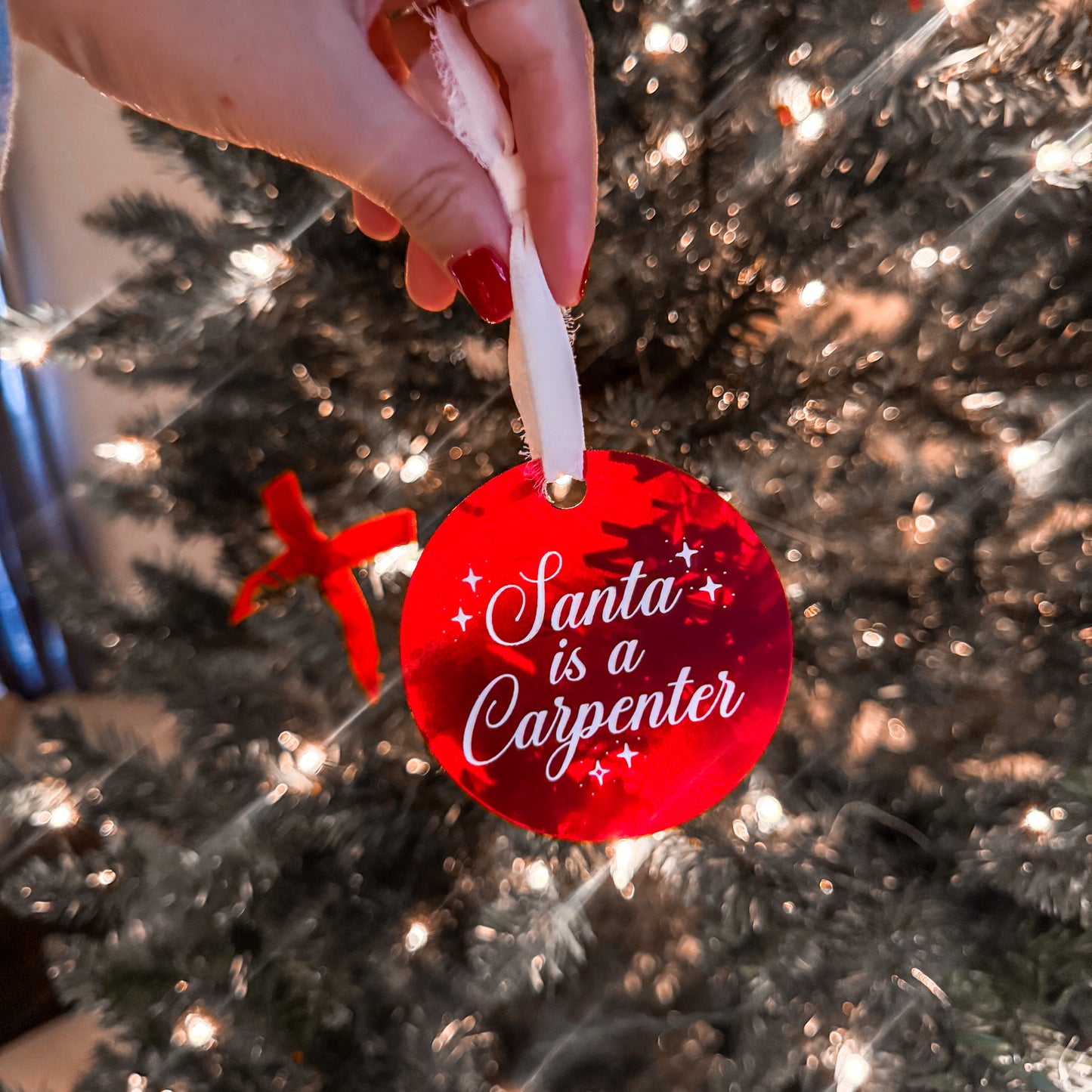 A red Sabrina Carpenter inspired Christmas ornament with the quote "Santa was a Carpenter" engraved and painted on the front and tied with a white chiffon ribbon.