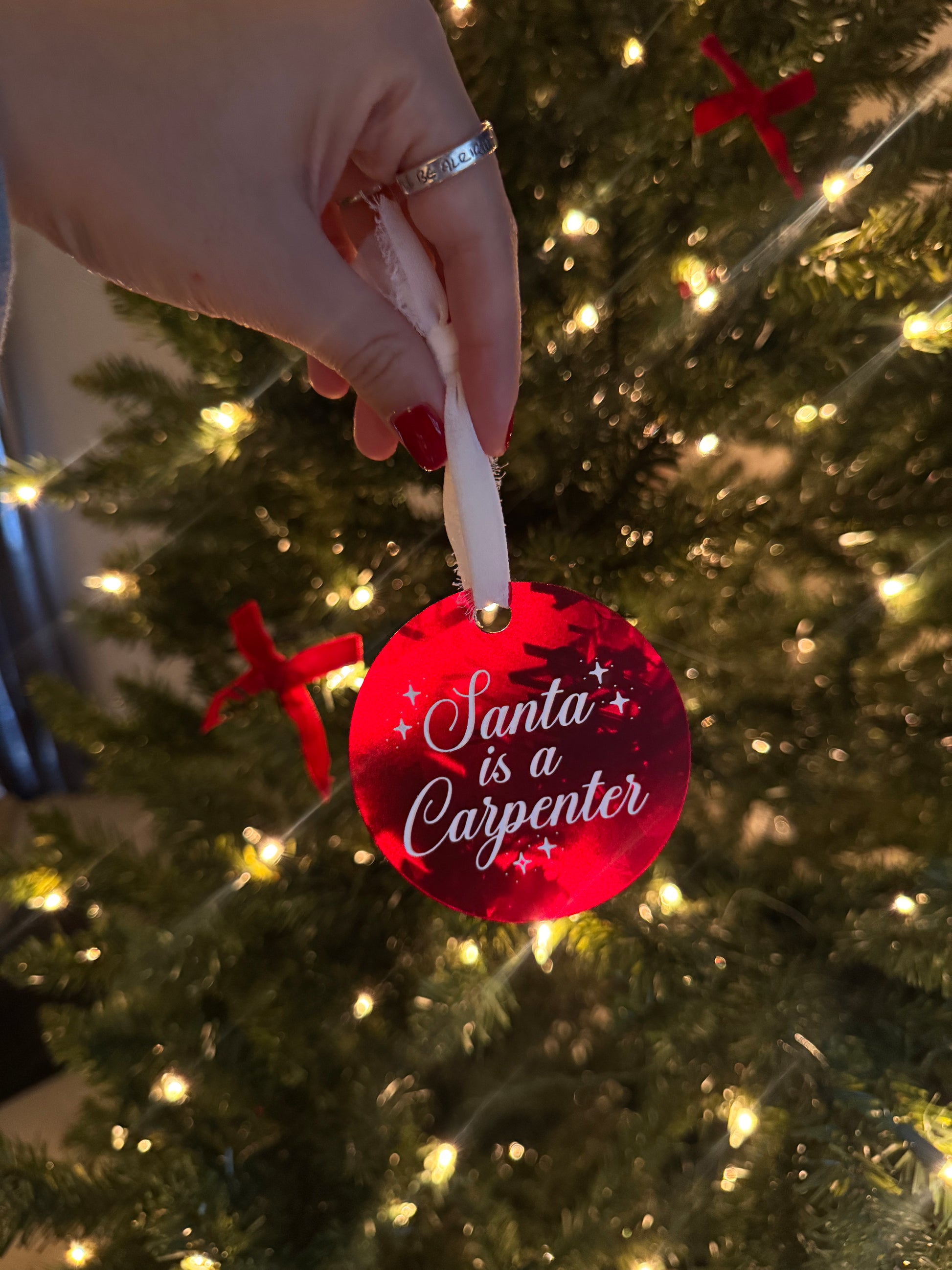 A red Sabrina Carpenter inspired Christmas ornament with the quote "Santa was a Carpenter" engraved and painted on the front and tied with a white chiffon ribbon.