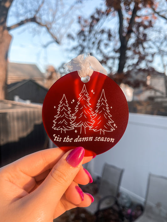 Round circle ornament made with frosted red acrylic, with a white chiffon ribbon tied at the top. Engraved and painted white on the front of the ornament are three Christmas trees, little sparkles, and the lyrics "'tis the damn season"