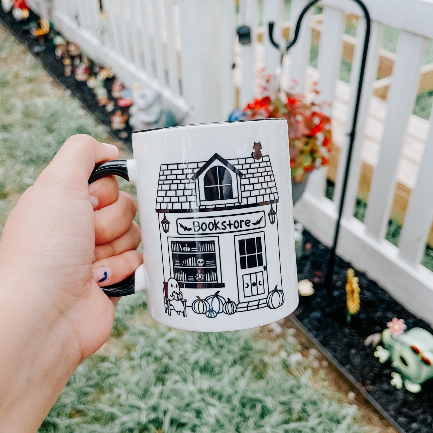 an 11 oz white mug with black handle and interior. the design feature a smile graphic of a spooky bookstore with a black cat sitting on top and a ghost reading a book in front.