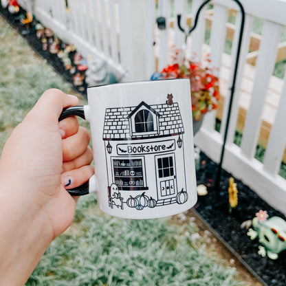 an 11 oz white mug with black handle and interior. the design feature a smile graphic of a spooky bookstore with a black cat sitting on top and a ghost reading a book in front.