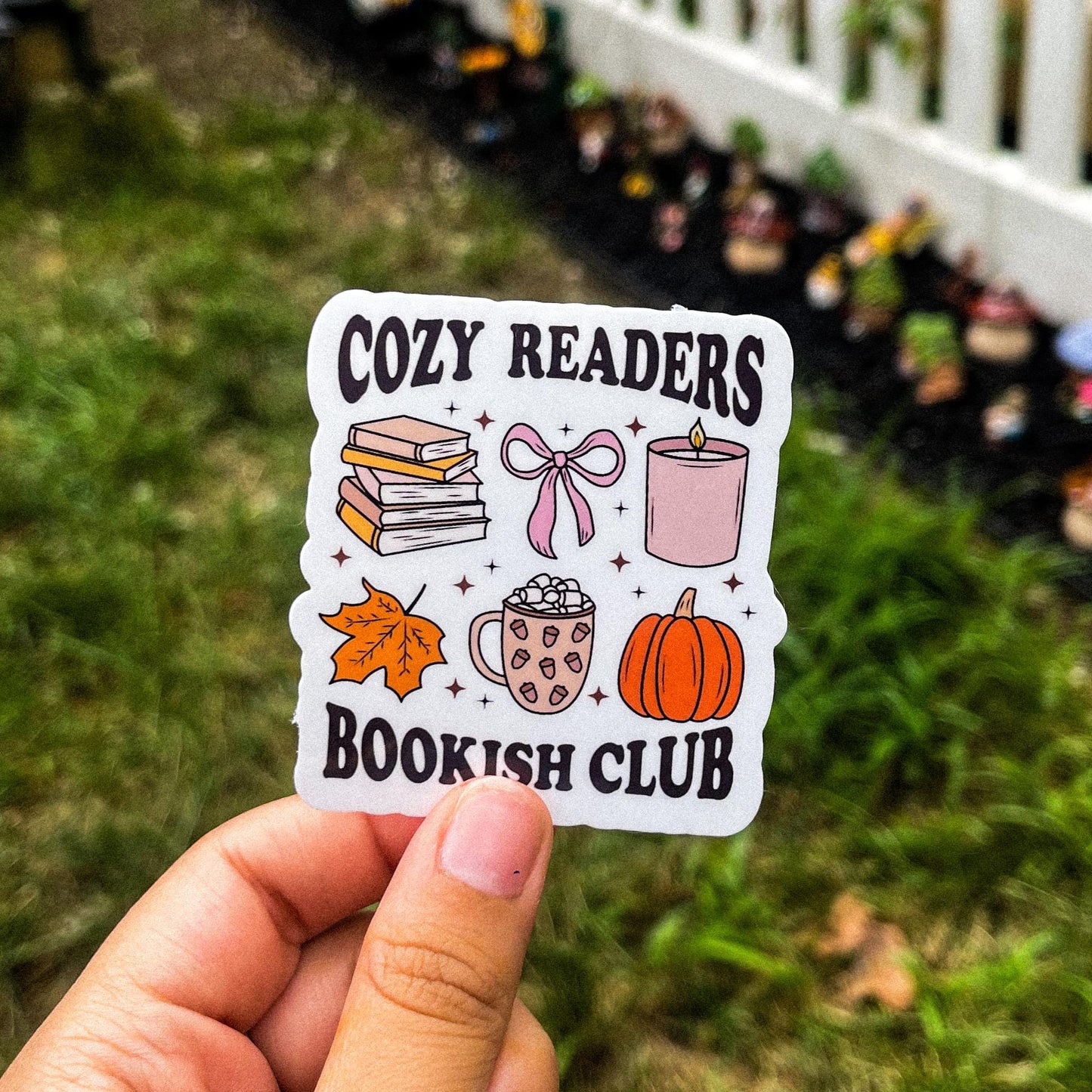 Bookish fall sticker with the words "cozy readers bookish club" in black. Between the words are different illustrations like hot chocolate, a pumpkin, a candle, a leaf, a bow, and a stack of books. 