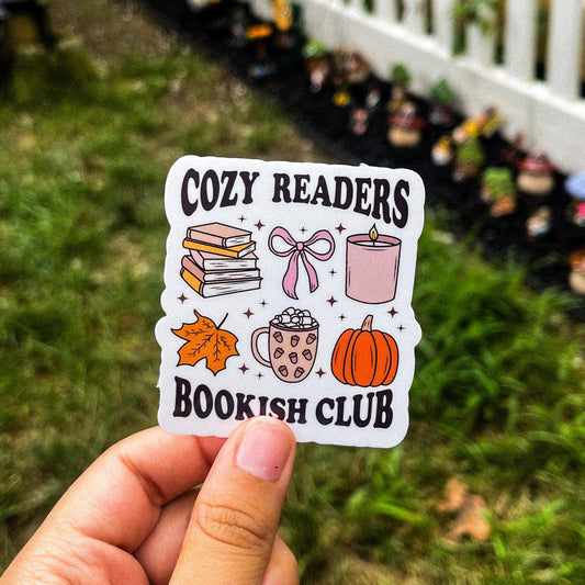 Bookish fall sticker with the words "cozy readers bookish club" in black. Between the words are different illustrations like hot chocolate, a pumpkin, a candle, a leaf, a bow, and a stack of books. 