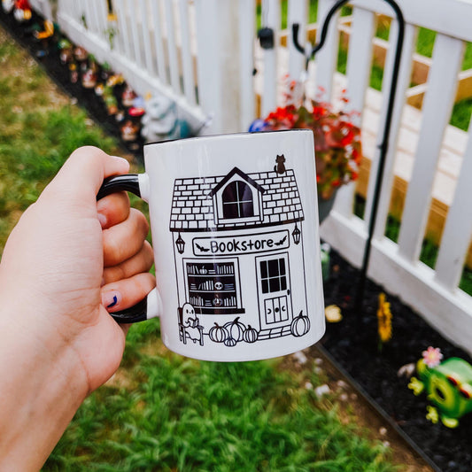 an 11 oz white mug with black handle and interior. the design feature a smile graphic of a spooky bookstore with a black cat sitting on top and a ghost reading a book in front.