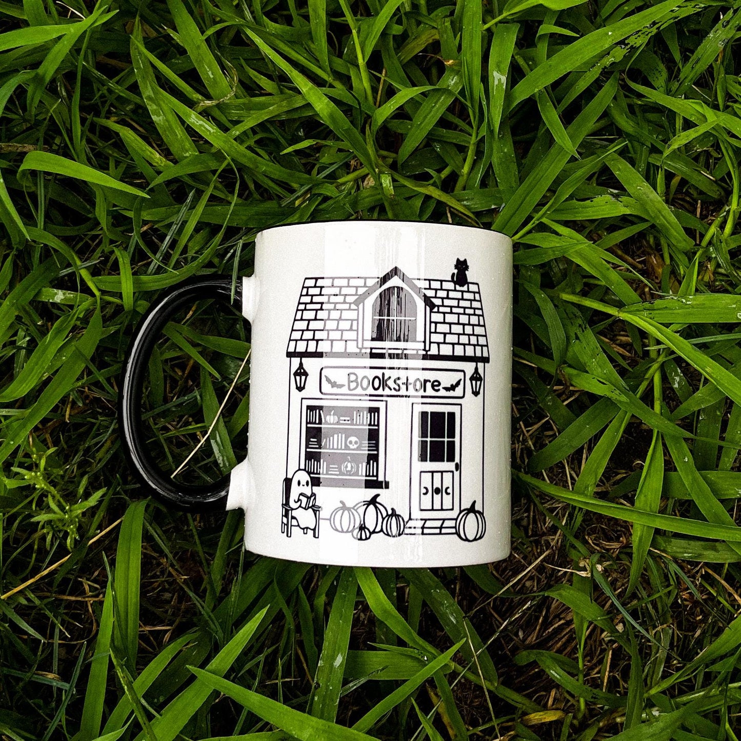 an 11 oz white mug with black handle and interior. the design feature a smile graphic of a spooky bookstore with a black cat sitting on top and a ghost reading a book in front.